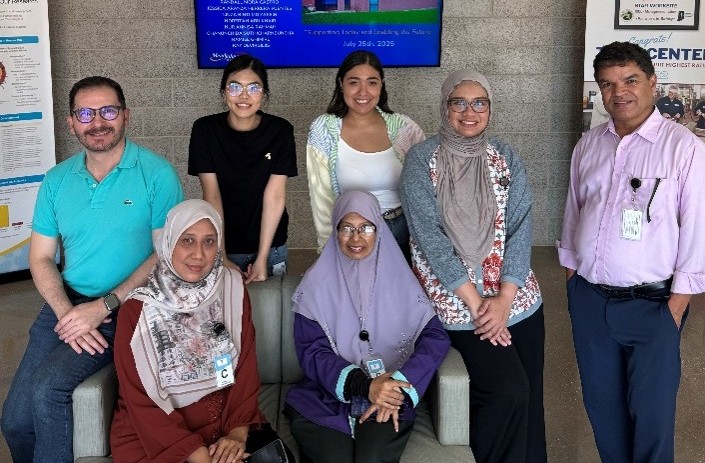 A group of seven program participants pose together indoors in front of informational display boards and a wall-mounted screen showing details about a food safety event. Three people are seated in front while four stand behind them. They are dressed in professional attire and appear to be at a training or program site.